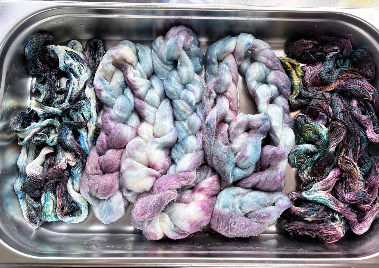 Dyed fiber and skeins of yarn in pink, blue, green, and dark shades in a metal tub.