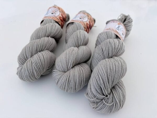 img_e119efb78c82.jpg Drie leigrijze strengen 4-ply garen met S7oRS Atelier-labels op een witte ondergrond.